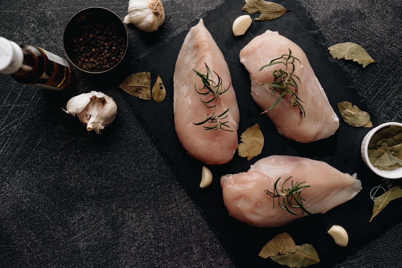 Raw chicken breasts with rosemary, bay leaves, and garlic on dark slate